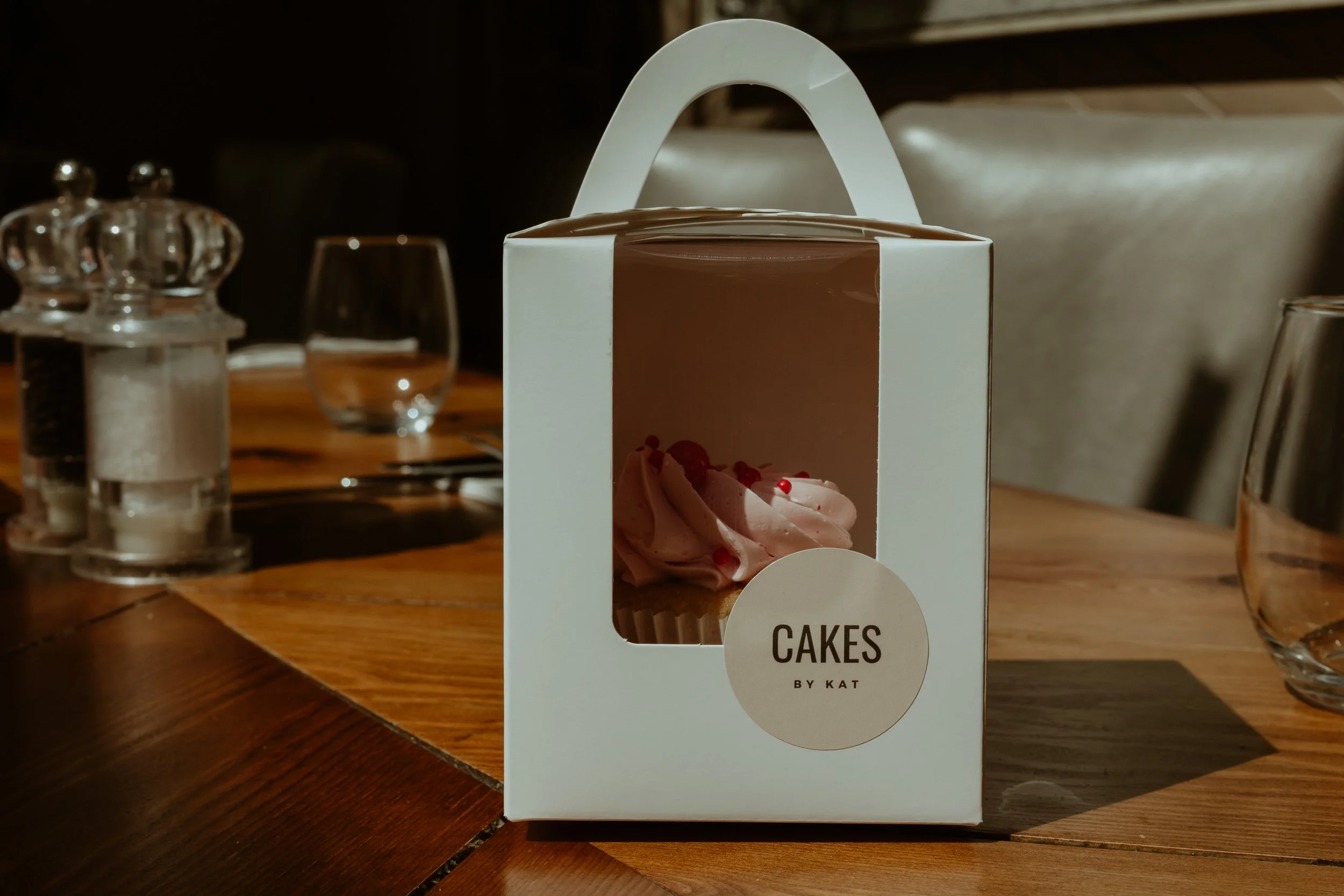 A pink frosted cupcake with red sprinkles inside a white takeout box with a clear window on a wooden table, with salt and pepper shakers, water glasses, and silverware in the background.