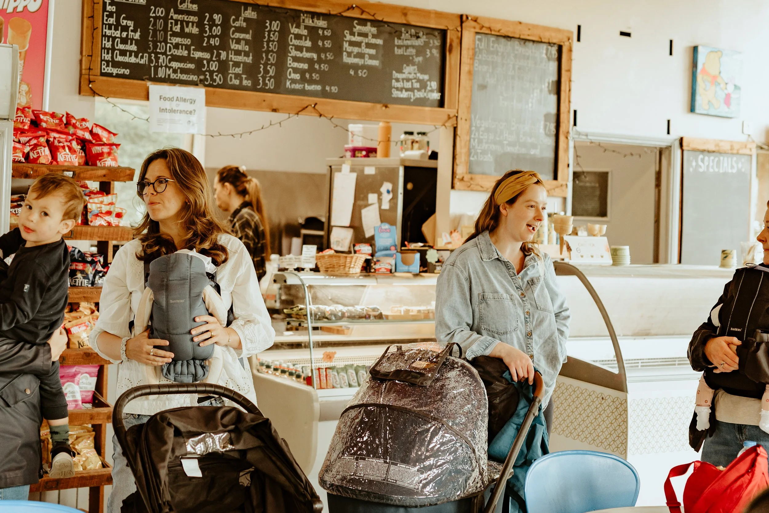 People inside a café or bakery, with a woman holding a backpack and a child, another woman behind the counter, and a person with a backpack and child on the right. The background shows menu boards, snacks, and a display case.