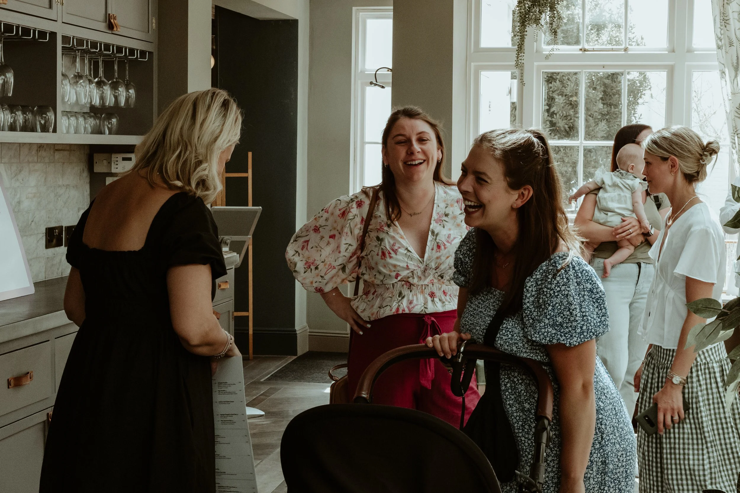 A group of women and a baby enjoying a cheerful moment at a social gathering inside a bright room with large windows, plants, and glassware.
