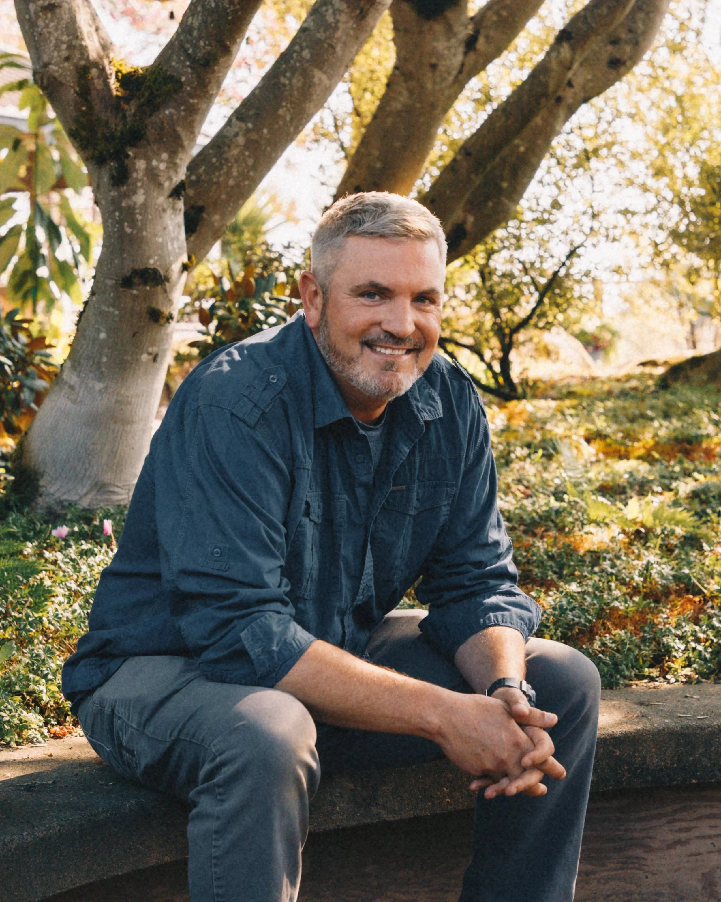 A man with gray hair and a beard smiling, seated outdoors in a park-like setting with trees and plants in the background, during daytime.