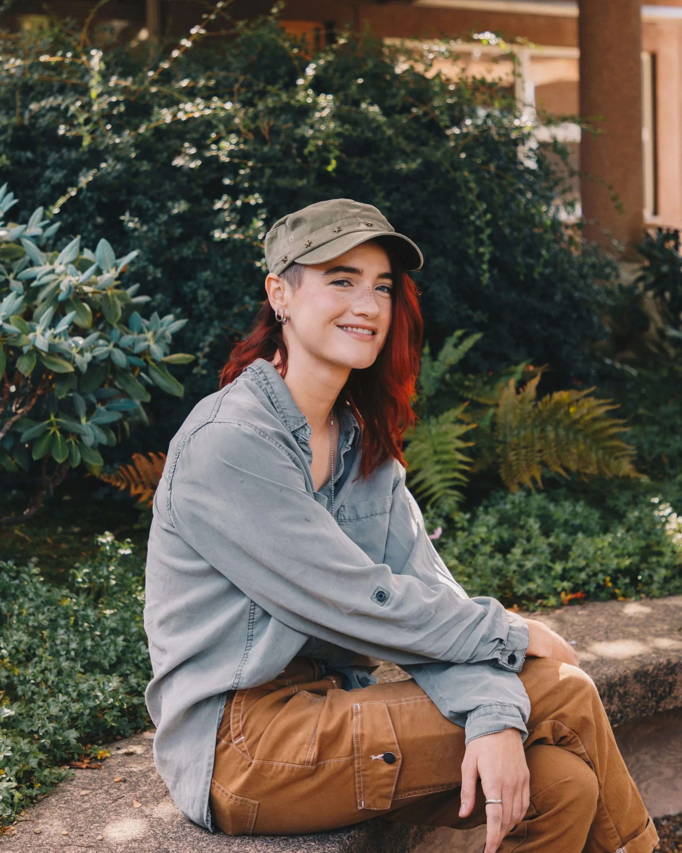 A young woman with red hair, freckles, and earrings, smiling while sitting outdoors on the ground near green bushy plants and a building, wearing a khaki cap, gray jacket, and brown pants.