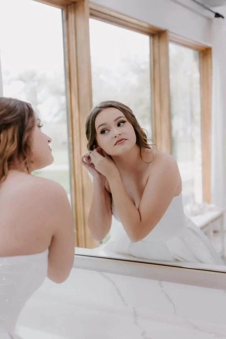 Young woman in a white strapless dress looking at herself in a mirror, adjusting her earring, in a room with large windows.