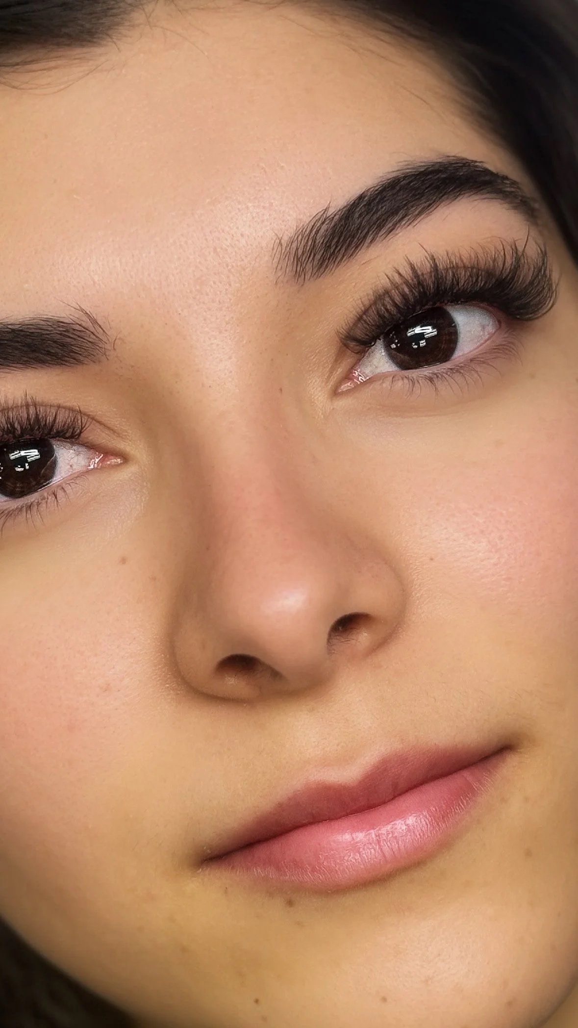 Close-up of a woman's face showing her eyes, nose, and lips with makeup including long eyelashes, well-groomed eyebrows, and subtle pink lipstick.