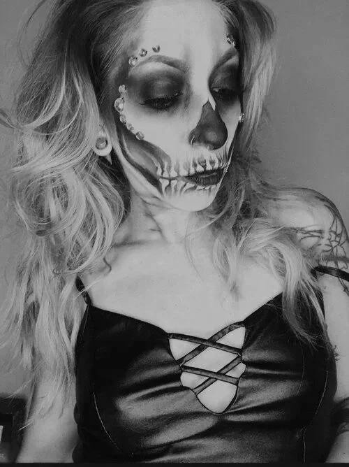 A woman with skull makeup on her face, wearing a black top with crisscross lacing in the front, and has curly blonde hair.