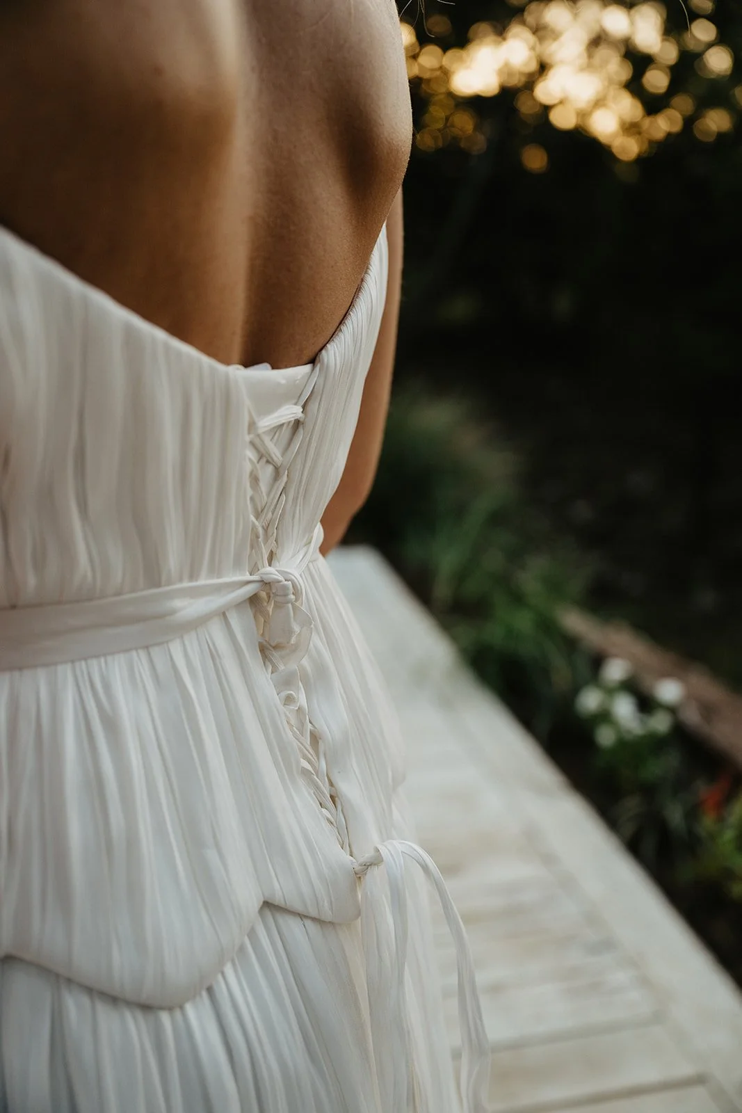 Close-up of the back of a woman wearing a white, pleated dress with a corset-style laced-up back, sitting outdoors at sunset.