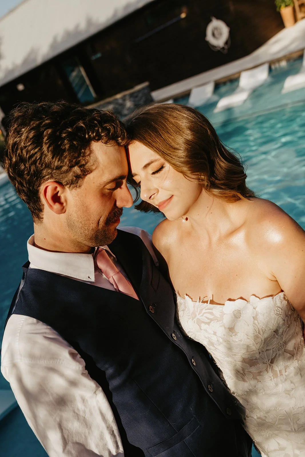 A man and a woman kiss each other near a swimming pool, with their foreheads touching and eyes closed, dressed in formal attire, with sunlight shining on them.