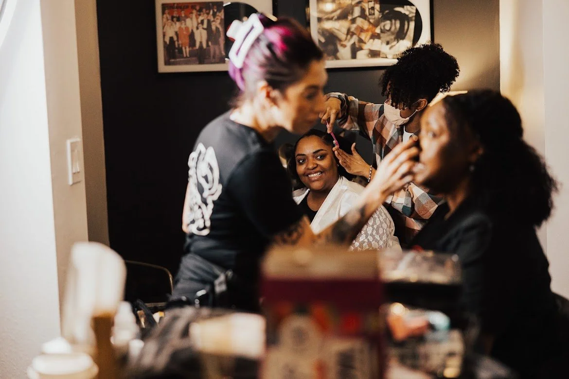A woman getting her hair styled by a hairstylist while two women sit nearby, smiling and talking in a salon or makeup room