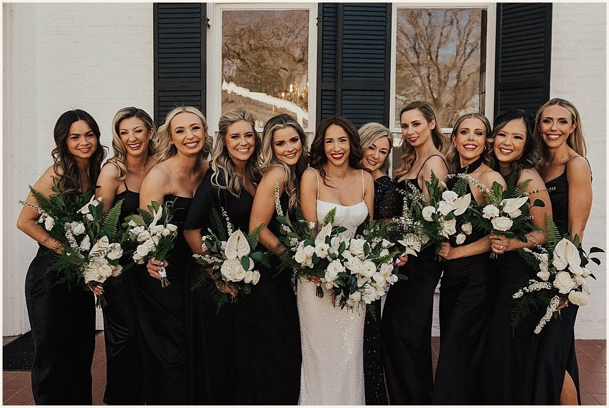 A group of women in black dresses, holding white floral bouquets, standing together outside a building with black shutters.