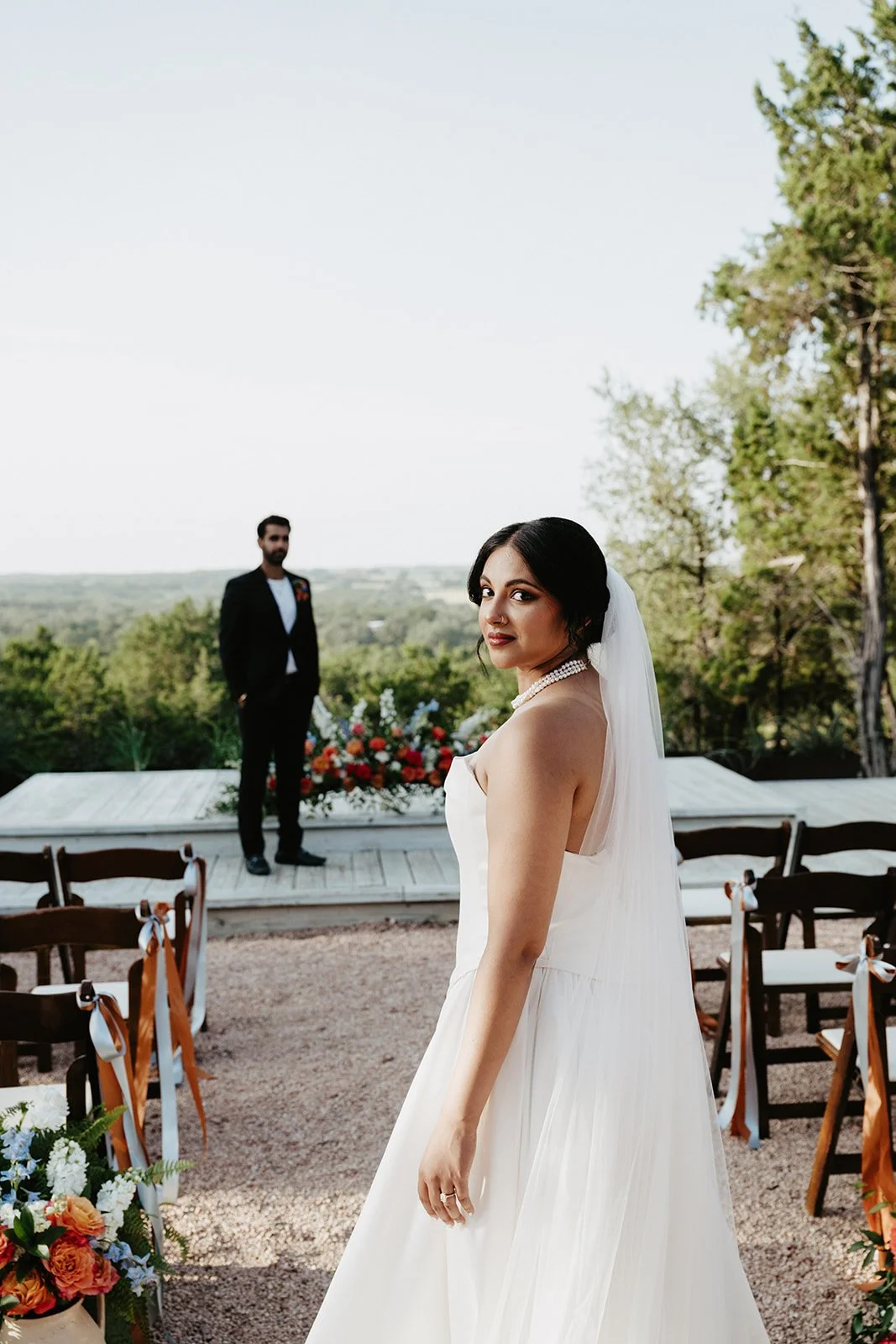 A bride in a white wedding dress stands outdoors during her wedding ceremony, with the groom in the background on a deck, surrounded by chairs decorated with ribbons and floral arrangements, against a backdrop of trees and open sky.