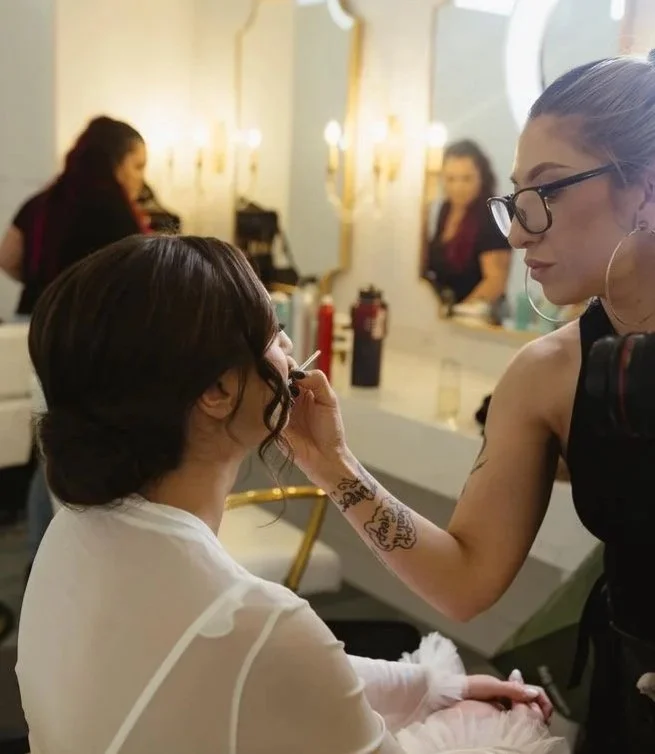 Makeup artist applying makeup to a seated woman in a salon with mirror and other clients in the background.