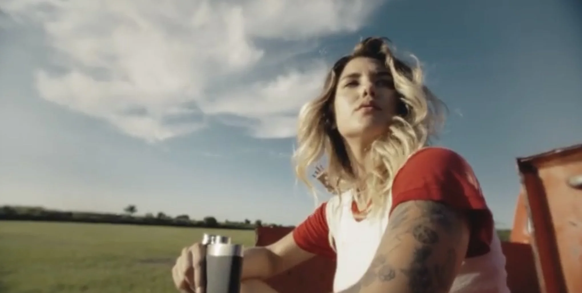 A woman with long, wavy blonde hair and tattoos on her arms sitting outdoors on a green field, holding a gun, with a blue sky and clouds in the background.