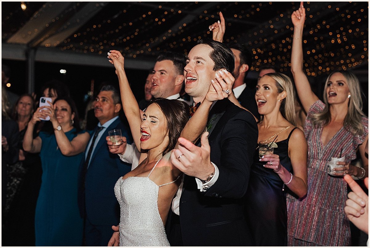 People dancing and celebrating at a wedding reception under string lights, with a bride and groom in the center enjoying the moment.