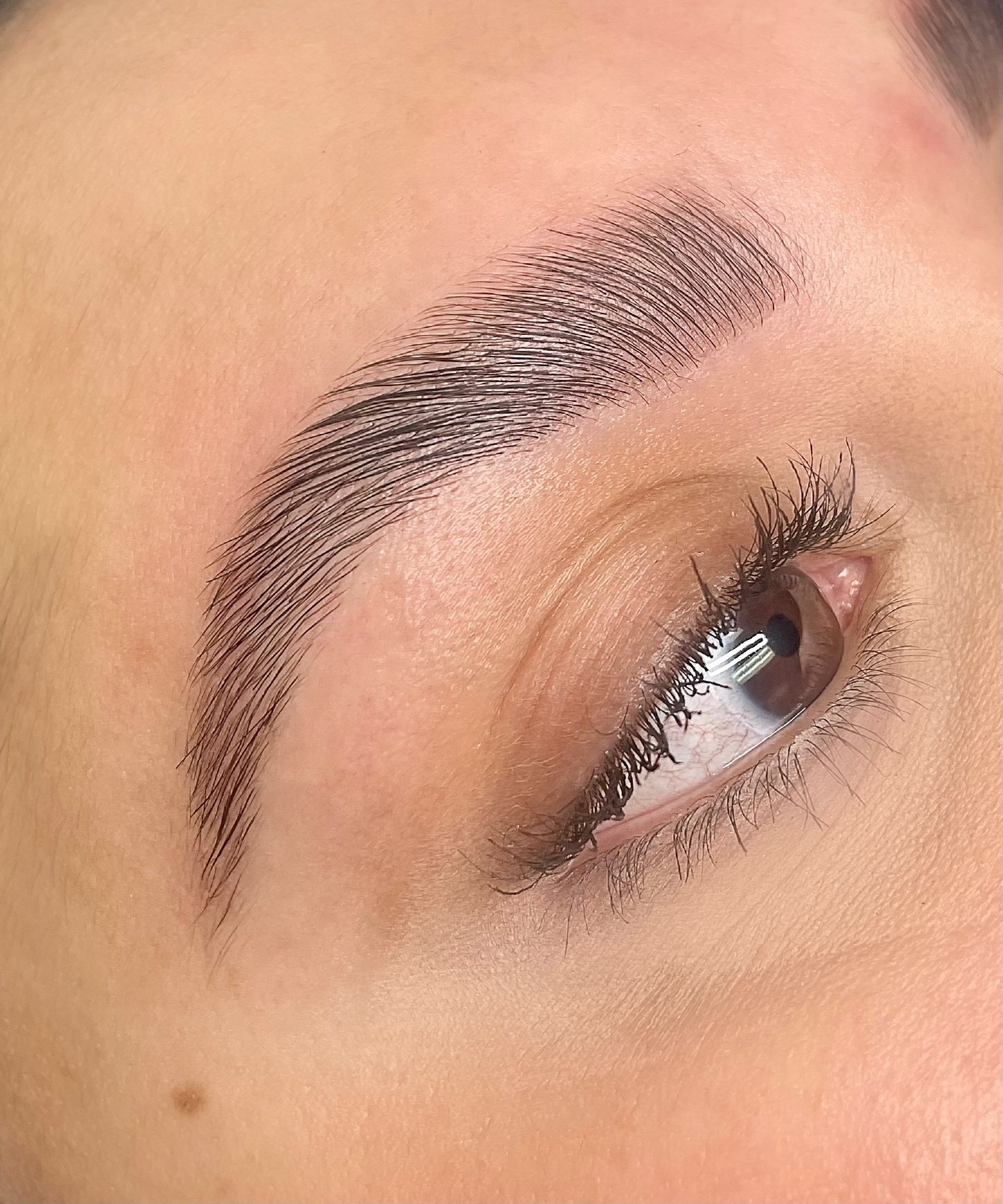 Close-up of a person's eyebrow, eyelash, and eye, with clear skin and a small mole on the cheek.