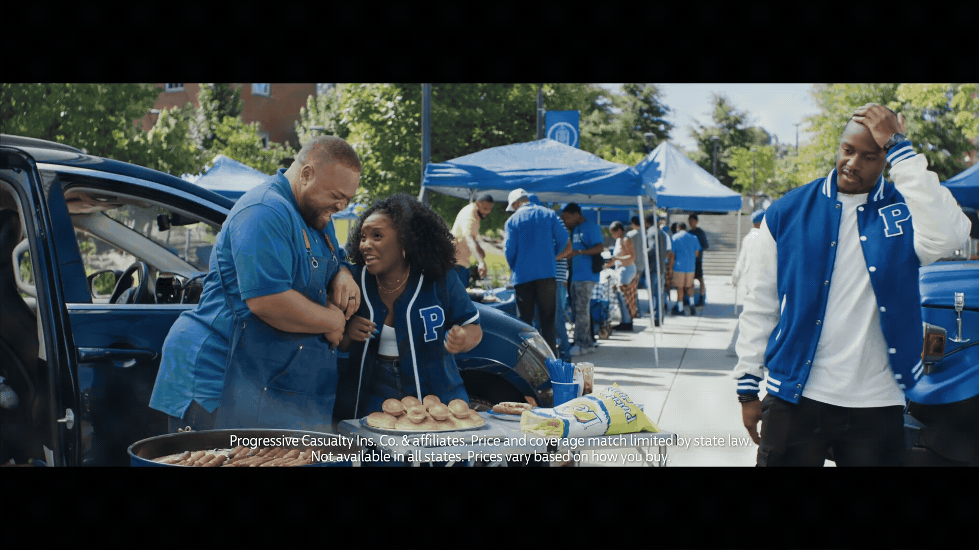 People at an outdoor barbecue ping pong event, wearing blue and white varsity jackets with a 'P'. They are near a blue vehicle and food table with hot dogs, buns, and condiments, under blue tents, surrounded by trees.