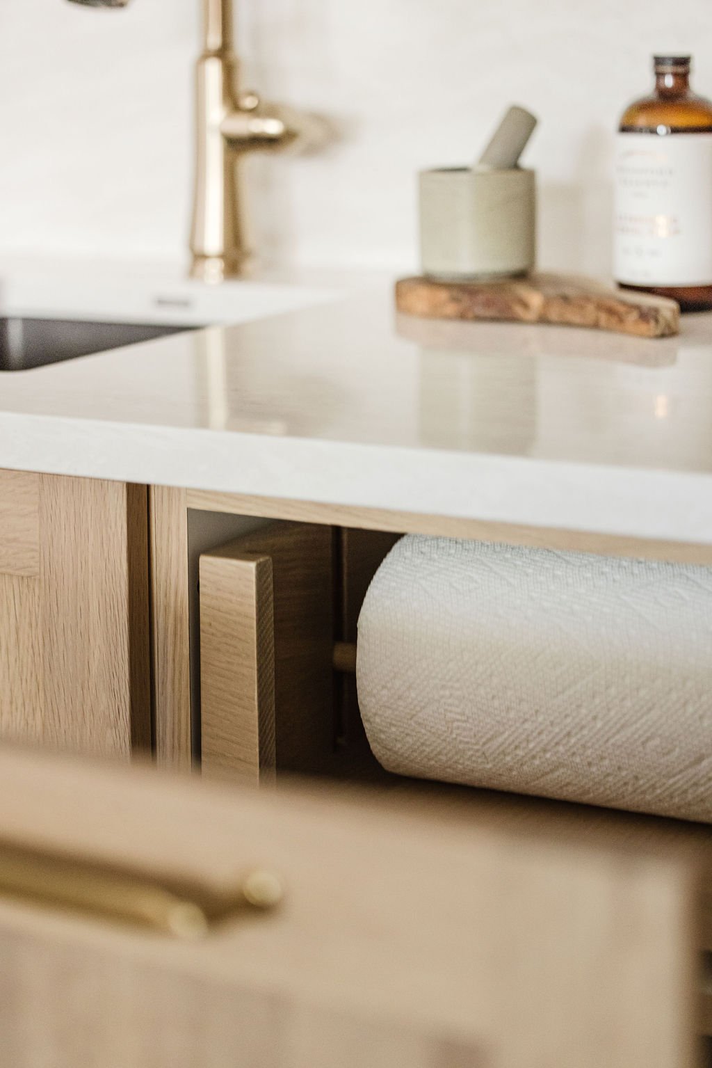 A detailed view of a custom light oak paper towel drawer, shown partially open to reveal the interior dowel and roll. The cabinetry features a flat-panel design and a sleek gold handle, with a white quartz countertop and gold gooseneck faucet visible