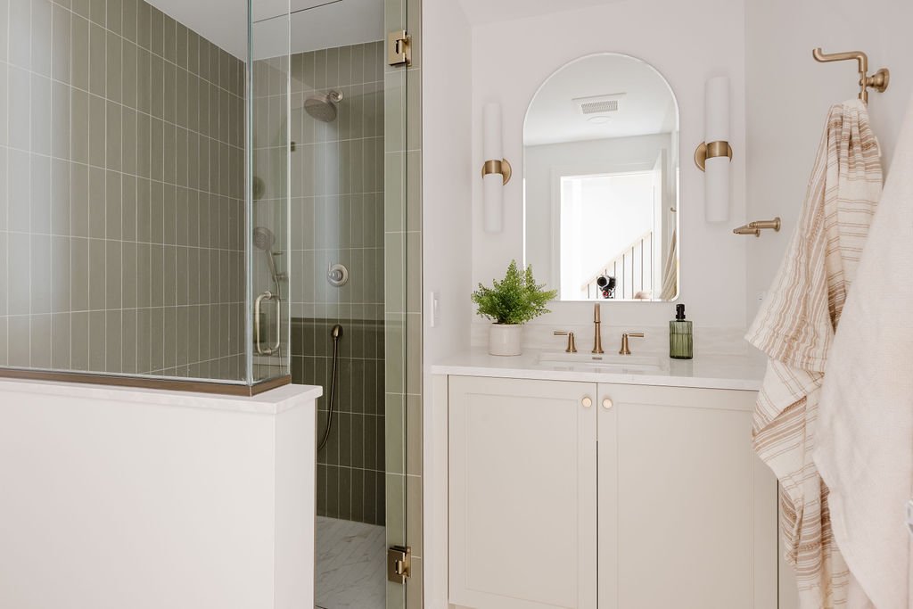 Modern bathroom featuring white shaker-style vanity, brass faucet and sconces, arched mirror, and glass shower with soft green tile walls.
