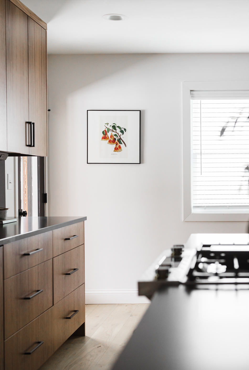Custom walnut kitchen cabinetry featuring a continuous vertical wood grain and minimalist matte black hardware. The design showcases flat-panel base drawers and upper cabinets set against a clean white wall with framed botanical artwork, emphasizing 