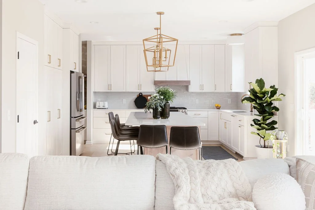 Luxury modern kitchen with white shaker cabinets, brass hardware, stainless steel appliances, a large island with black barstools, light hardwood floors, and a gold geometric chandelier in an open-concept living space.
