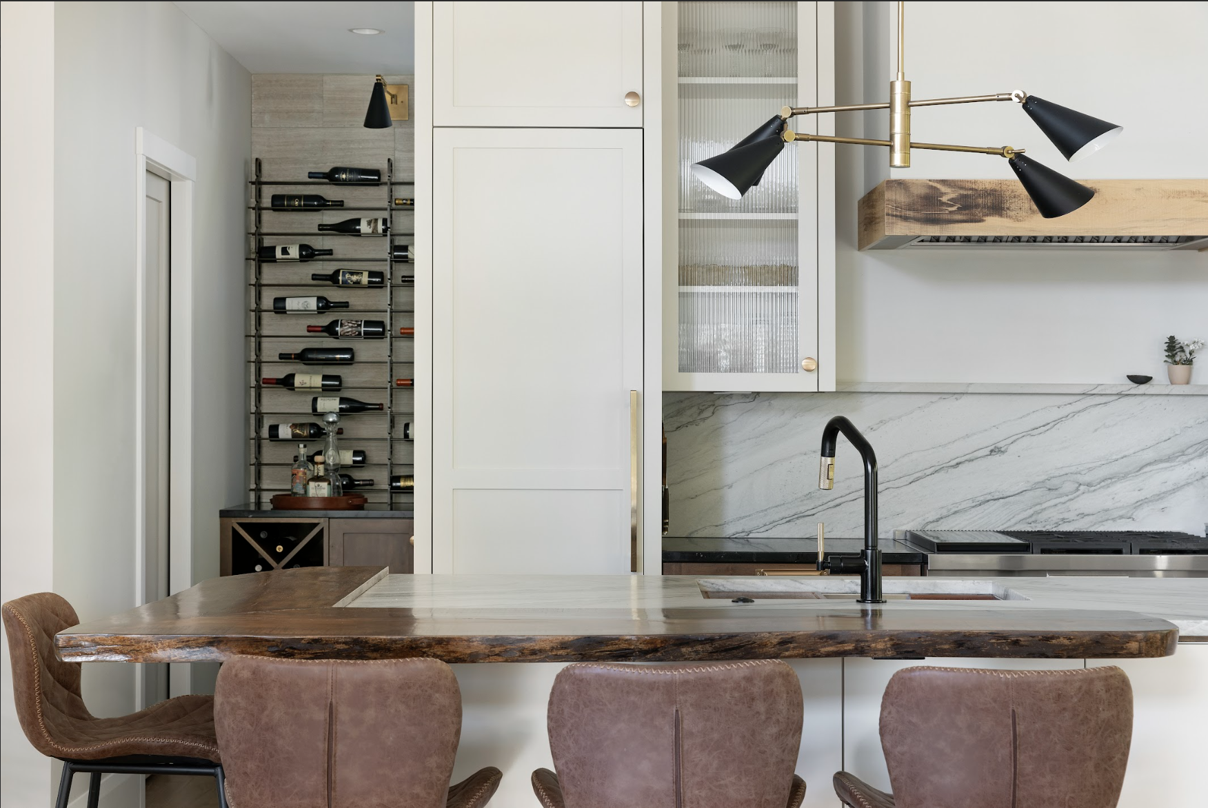 Custom white kitchen cabinetry with integrated wine storage, wood range hood, marble backsplash, and live-edge island countertop.