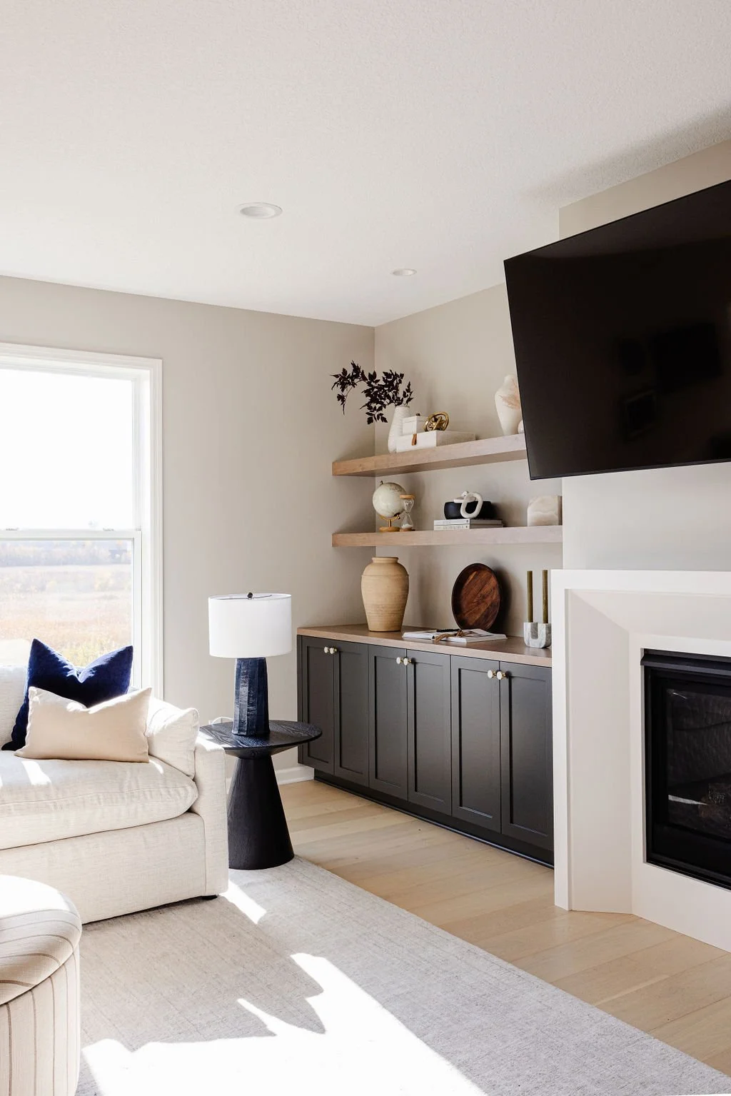 Bright modern living room featuring black built-in cabinets, floating wood shelves, neutral decor accents, and a mounted TV beside a fireplace.