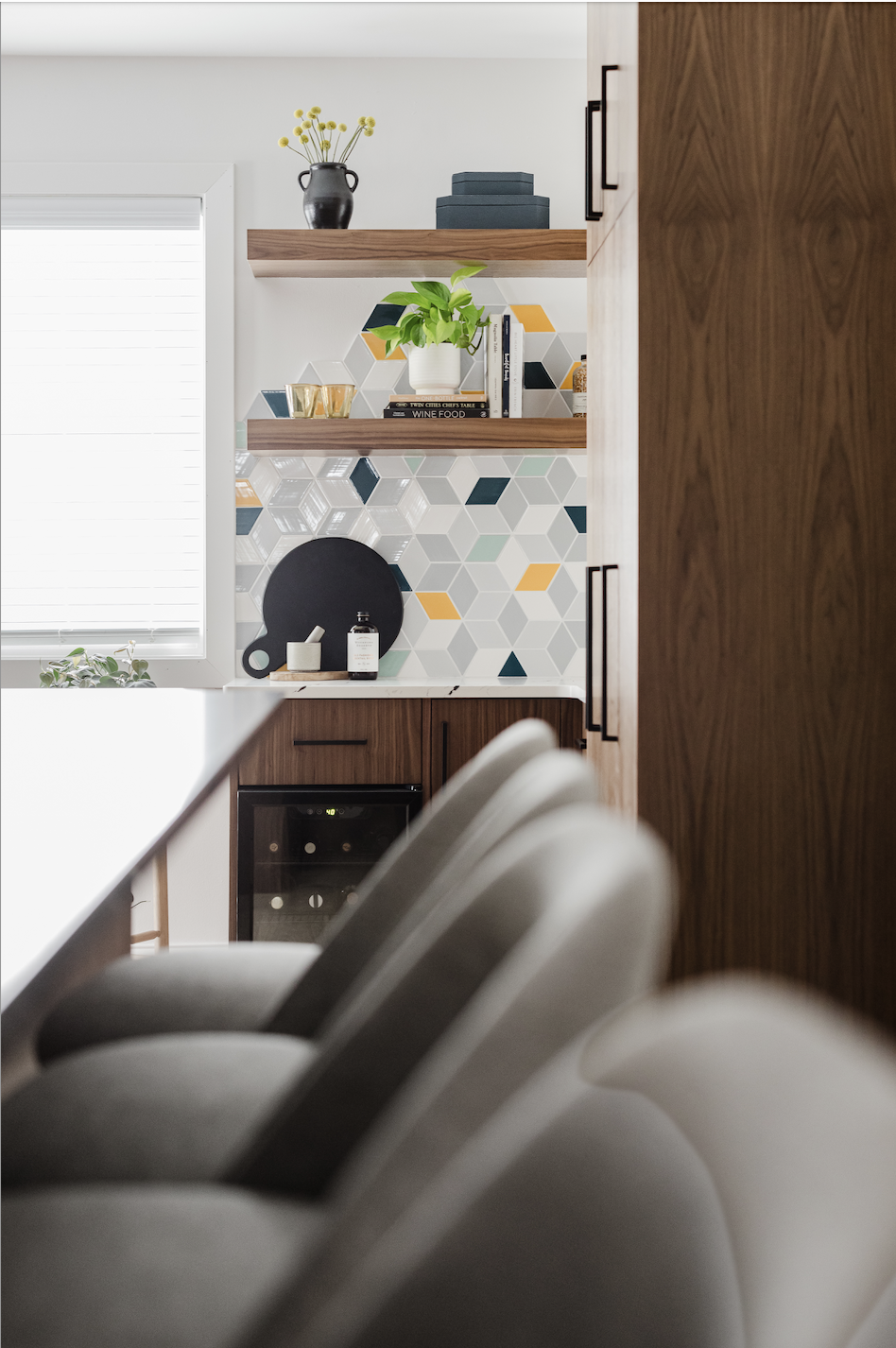 Perspective view looking past grey barstools toward a custom integrated walnut beverage station. This minimalist modern design showcases bookmatched vertical wood grain cabinetry, colorful geometric cube-patterned tile, and custom-matched floating sh