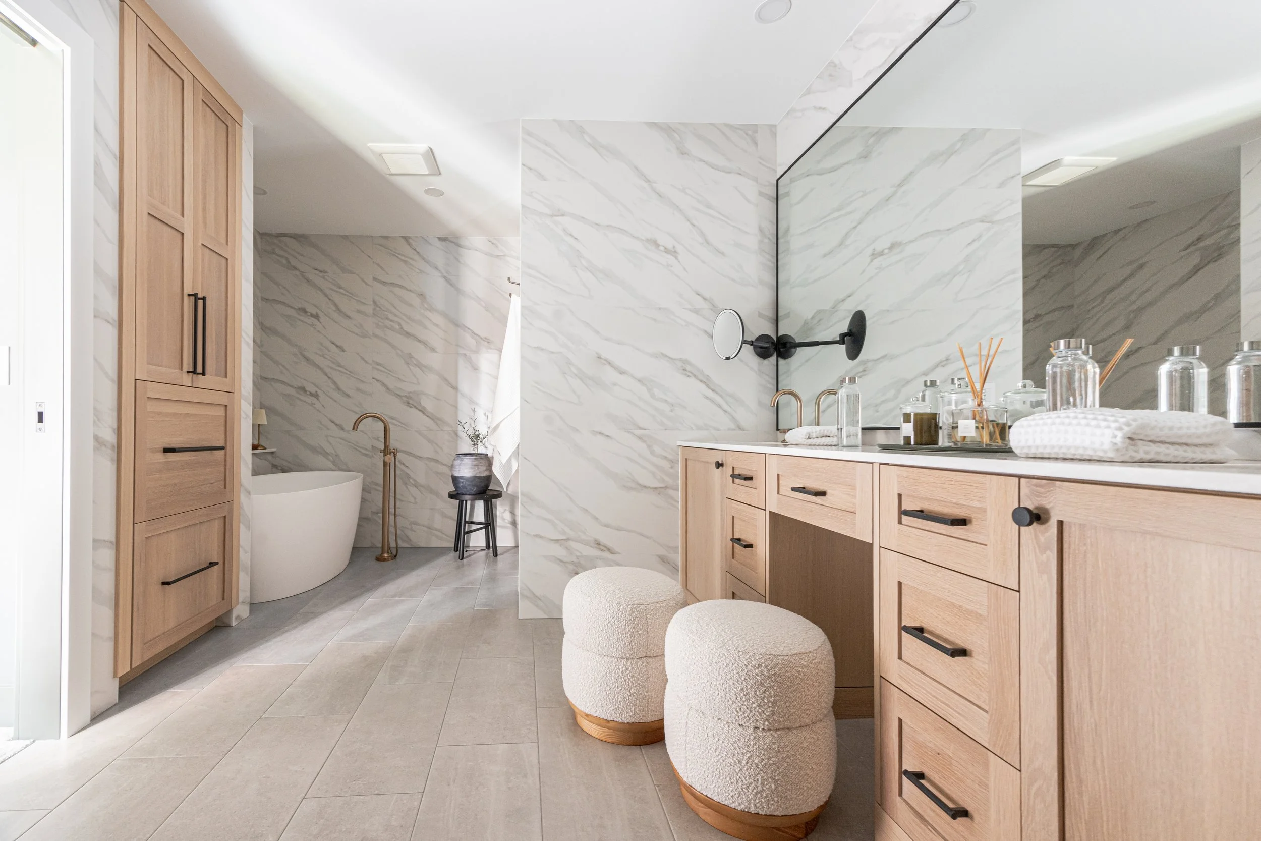 Light wood cabinets in a spa-style bathroom with marble walls, a double vanity, and a freestanding tub nearby.