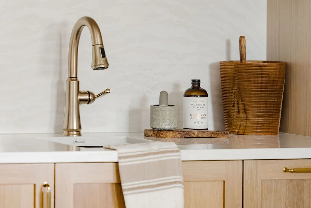 Detail shot of a light oak wet bar featuring a gold gooseneck faucet and a white marble-patterned backsplash. A wooden ice bucket and cocktail ingredients sit on the white countertop above flat-panel wood cabinetry.