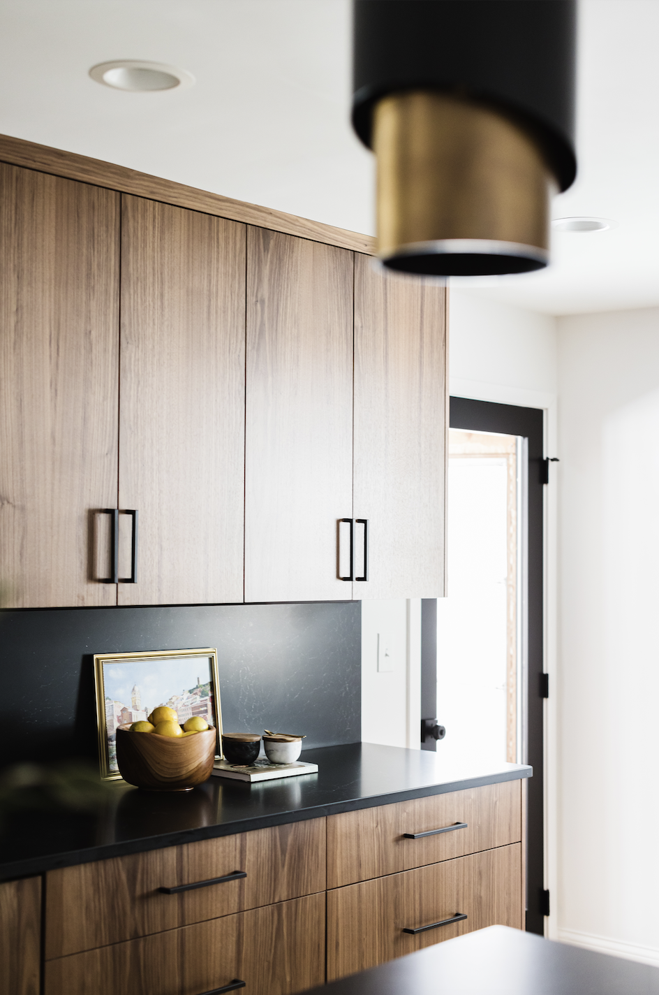 Modern walnut upper and lower cabinetry featuring slab-style doors, a matte black backsplash, and slim black pulls in a bright, contemporary kitchen.