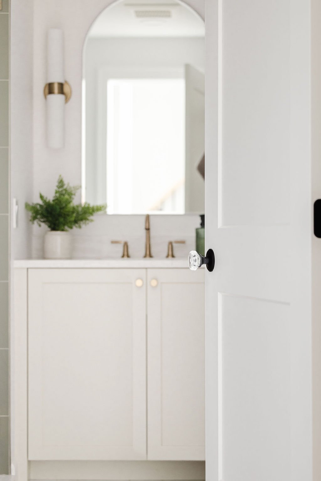 View through an open door with a crystal knob of a white shaker bathroom vanity, arched mirror, and brass fixtures.