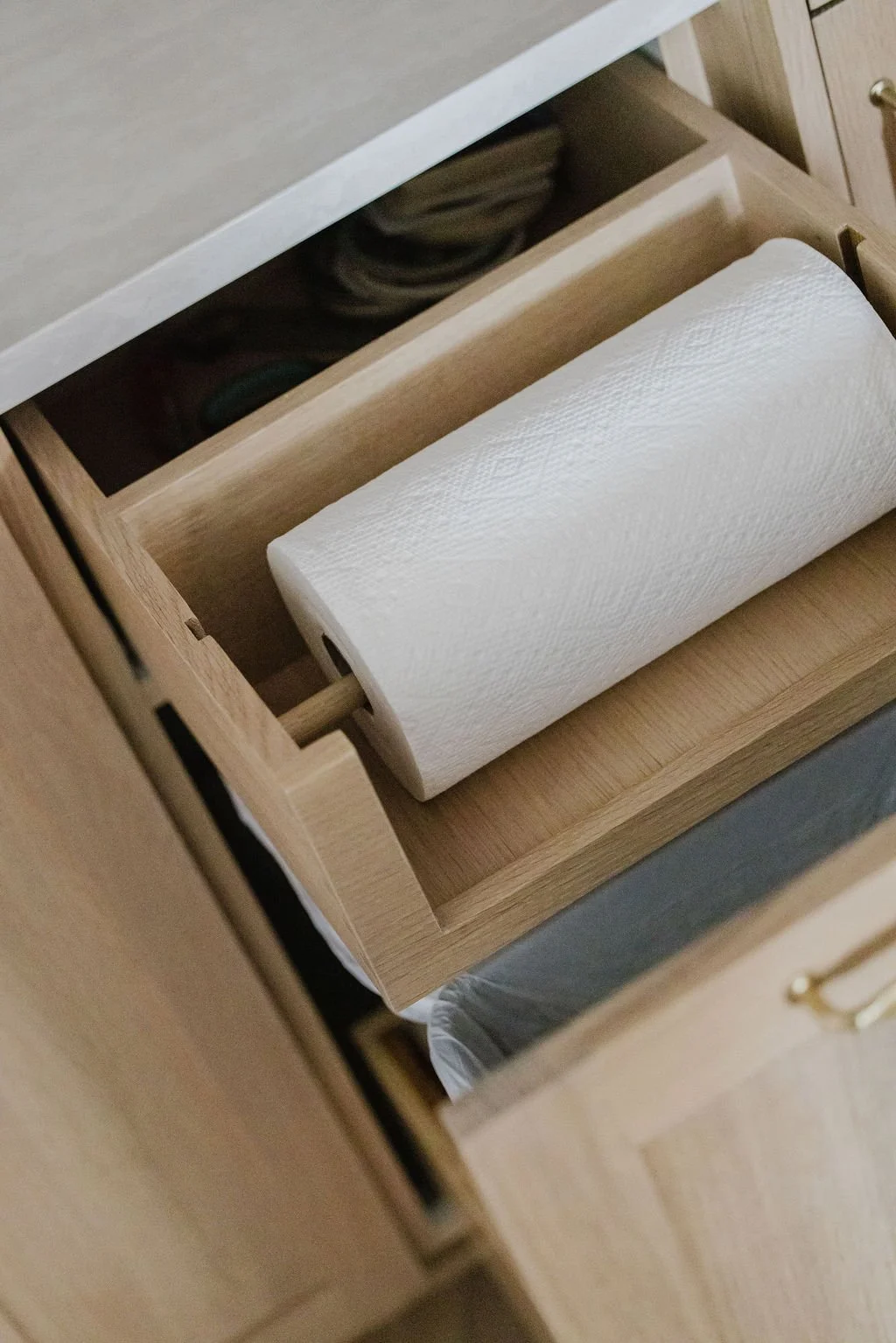 High-angle detail of a custom light oak pull-out cabinet drawer. The top compartment features an integrated wooden dowel holding a paper towel roll, while the bottom section reveals a pull-out waste bin, showcasing a space-saving and functional cabin