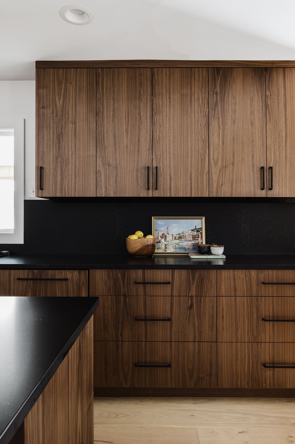 Modern kitchen featuring custom walnut upper and lower slab cabinetry with matte black hardware, a fruit bowl, a painting with a gold frame, dark countertops, and a minimalist black backsplash.