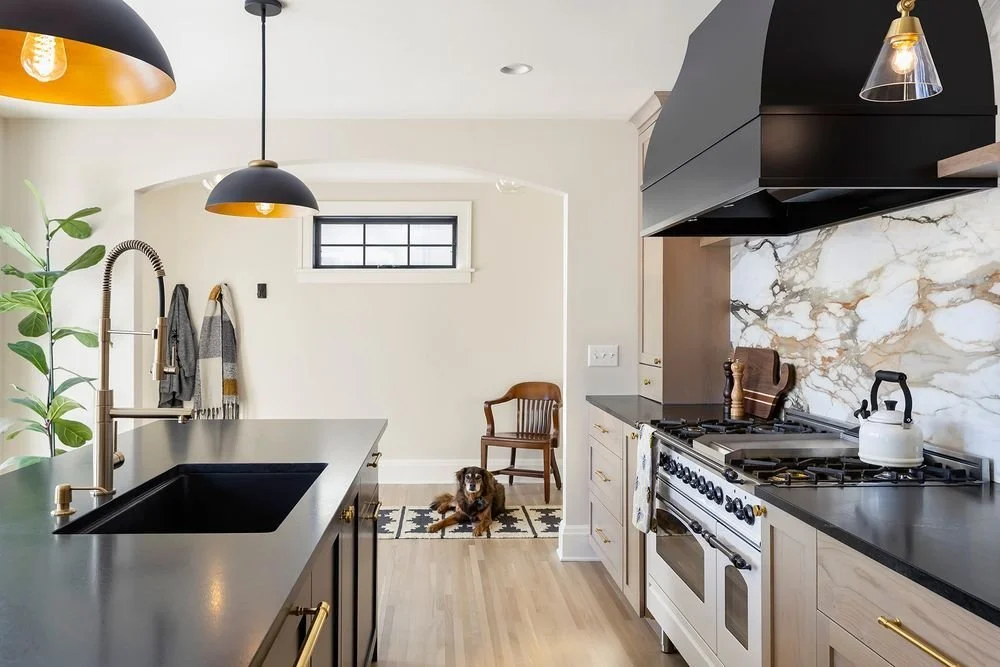 Two-tone kitchen design featuring natural wood cabinets, black range hood, marble backsplash, and oversized black pendant lighting. A dog lounges in the background.