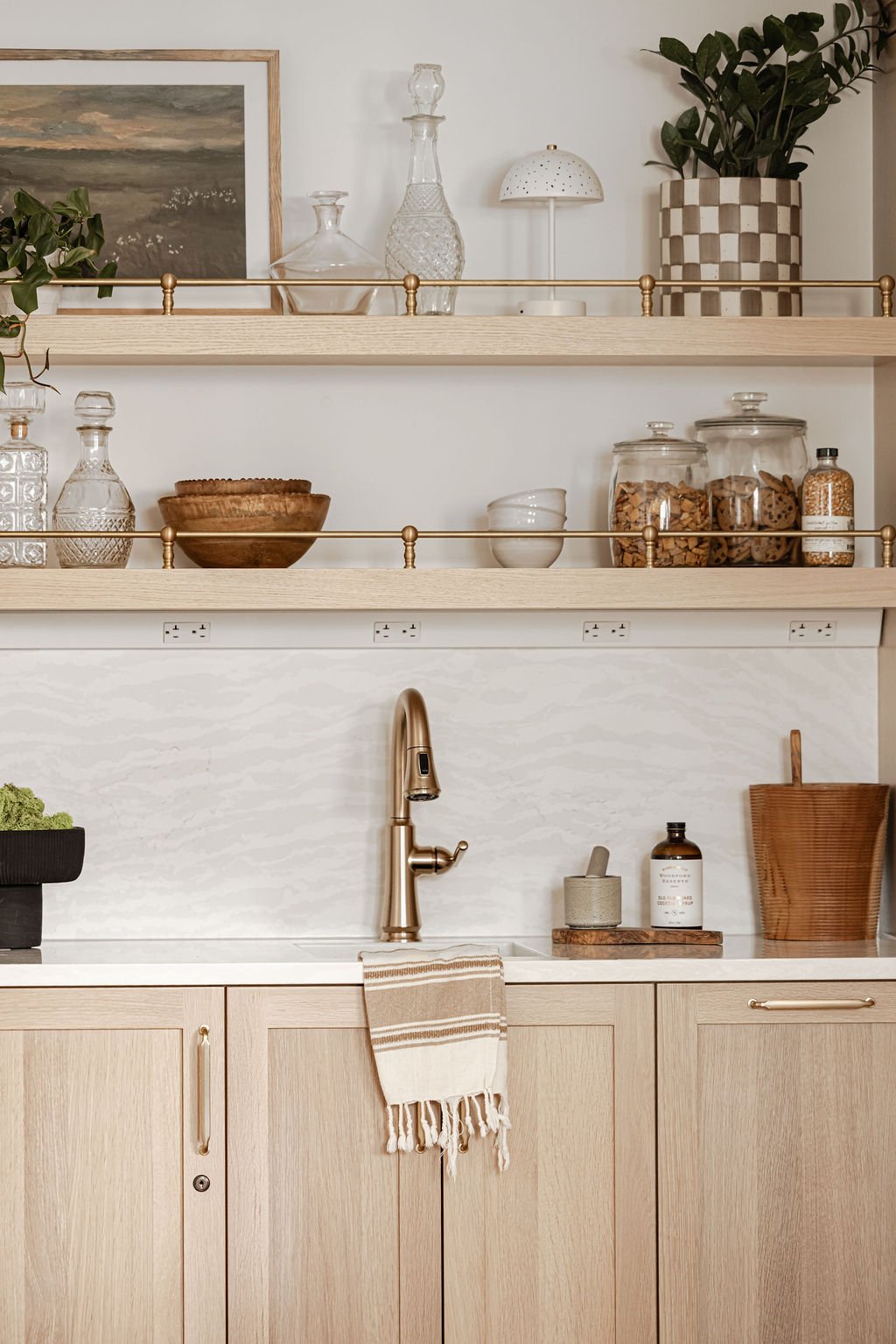 Detail shot of a light oak wet bar featuring flat-panel cabinets and two floating shelves with decorative brass gallery rails. A gold gooseneck faucet is centered against a white marble-patterned backsplash with subtle veining.