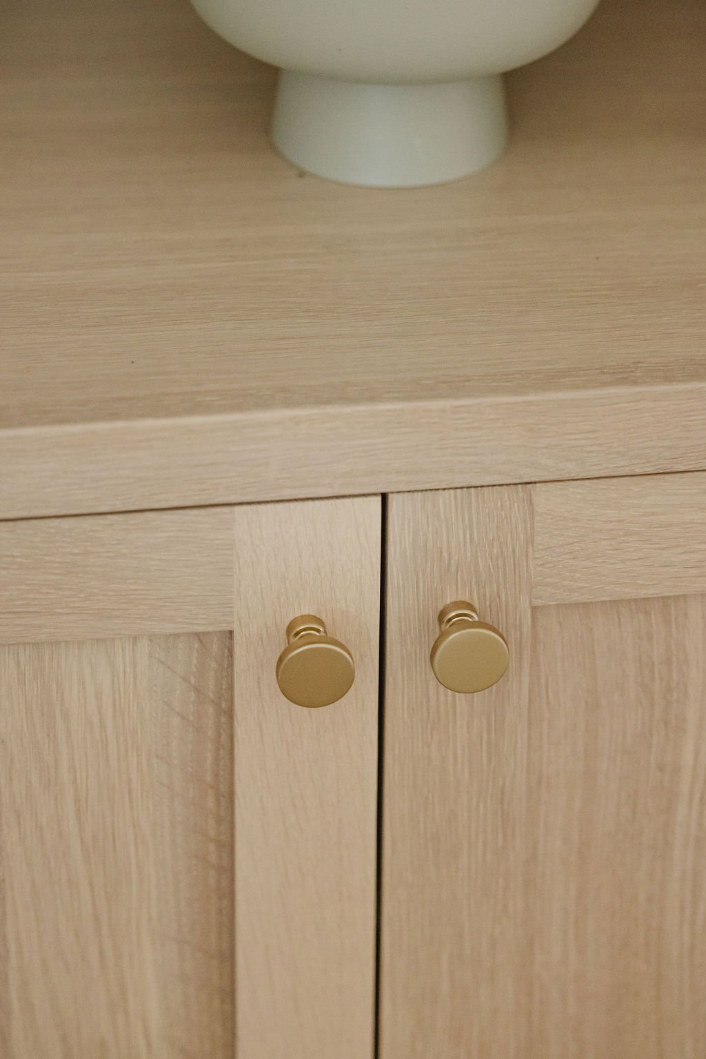 Bright bathroom corner featuring a white shaker vanity with gold knobs and marble countertops. Two neutral-toned fringed towels hang from brass hooks on a clean white wall next to a modern tube sconce.