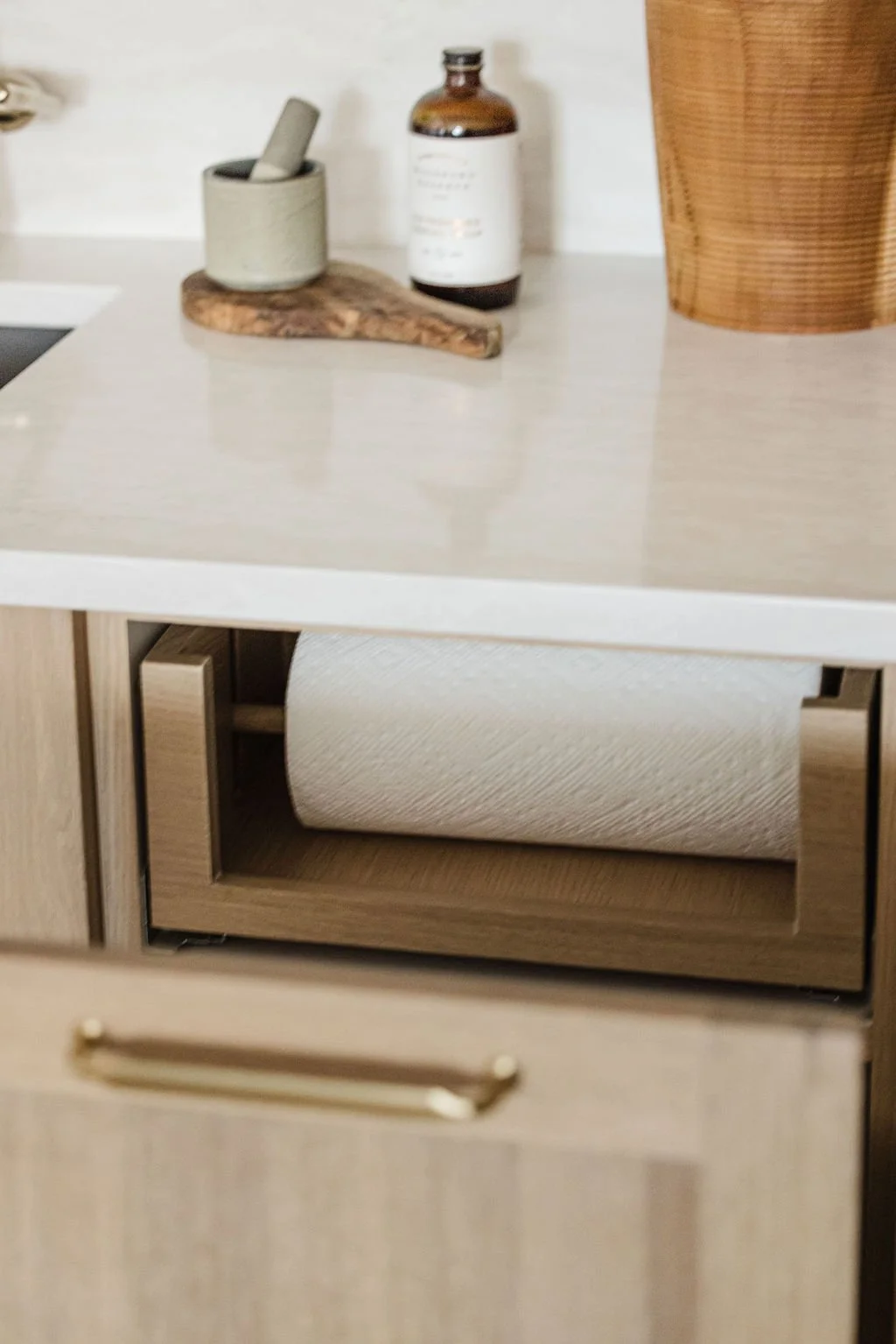 Interior detail of a custom light oak pull-out drawer showing a wooden paper towel holder. The drawer side features high-quality dovetail joinery and a laser-engraved "Sean's Cabinetry" logo. In the soft-focus background, a gold gooseneck faucet stan