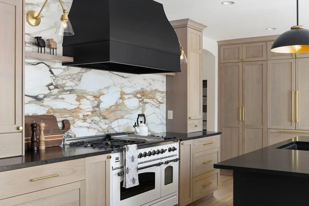 Custom kitchen cabinetry in natural light wood with brass hardware, paired with a statement marble backsplash, white professional-style range, and bold black vent hood surround.