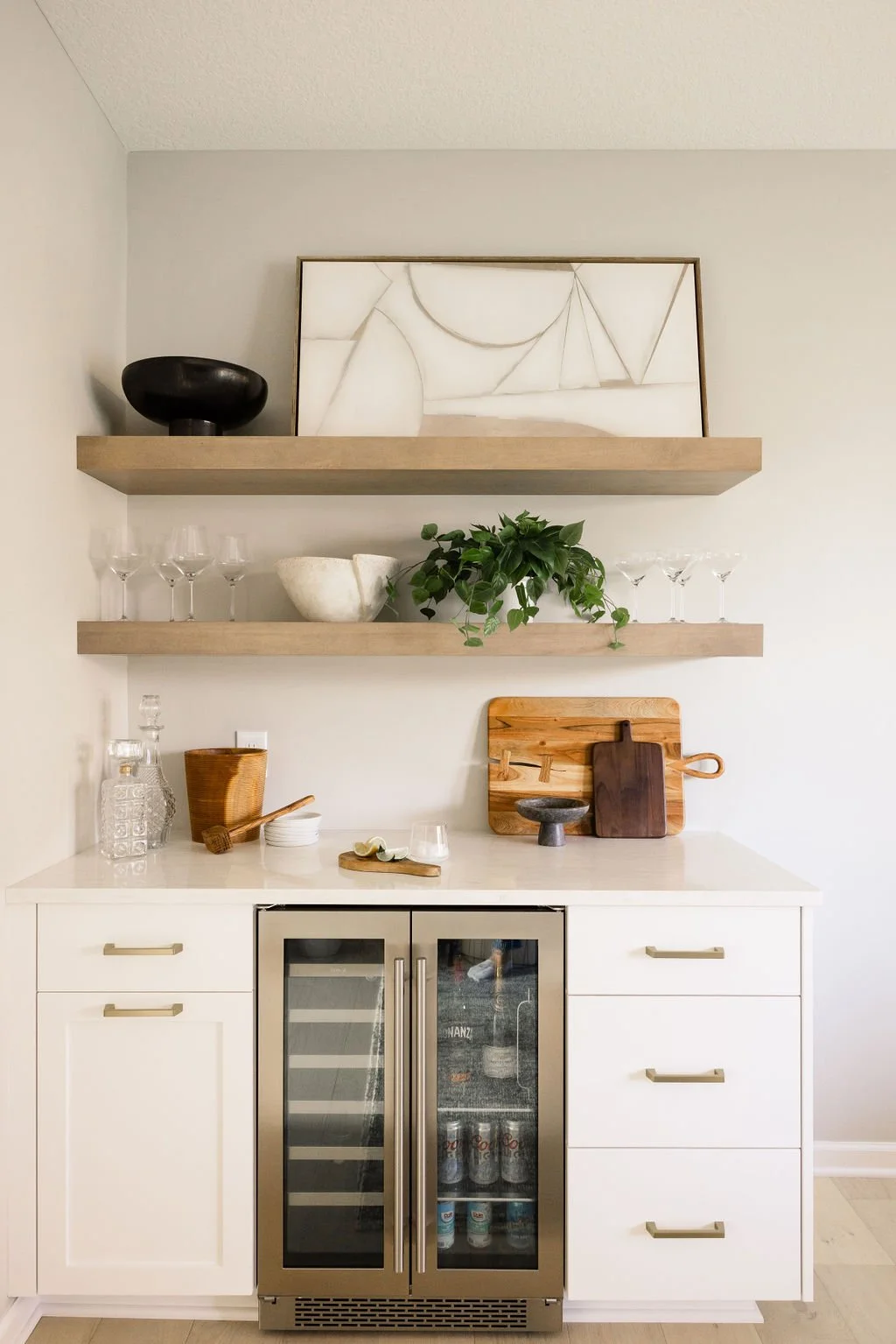 Modern home bar featuring custom white shaker cabinets, wood floating shelves, and an integrated stainless steel beverage fridge, styled with glassware and decor.