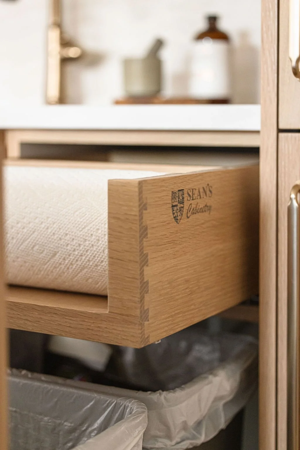 Close-up of a custom light oak pull-out drawer with dovetail joinery, featuring a laser-engraved "Sean's Cabinetry" logo on the side. The drawer is designed to hold a paper towel roll and is positioned above an integrated waste bin.