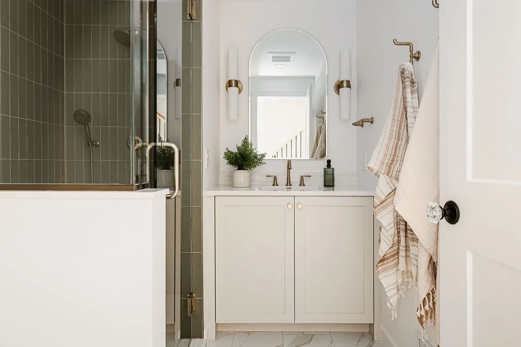 Bright bathroom featuring white shaker-style cabinets, brass faucet and sconces, arched mirror, and tiled glass shower enclosure.