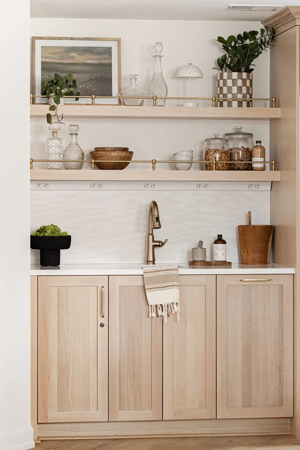 Custom light oak wet bar featuring flat-panel cabinets and two floating shelves with decorative brass gallery rails. The setup includes a white marble backsplash, a gold faucet, and curated barware decor.