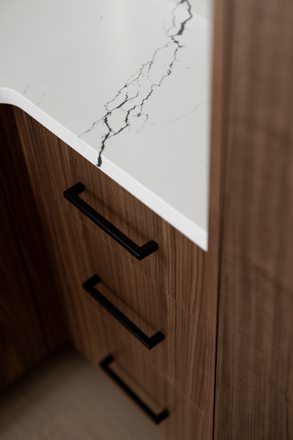 Close-up detail of high-end walnut cabinetry craftsmanship featuring bookmatched vertical grain and matte black linear pulls. The cabinets are paired with a luxury white quartz countertop with dramatic black veining, showcasing precise millwork and c