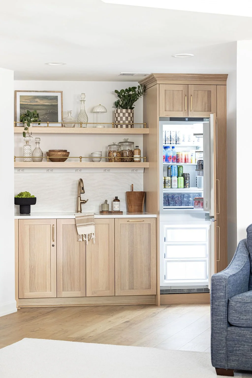 A vertical view of the custom light oak wet bar featuring an open integrated refrigerator and freezer unit. Complete with flat-panel lower cabinets and floating shelves with brass gallery rails.