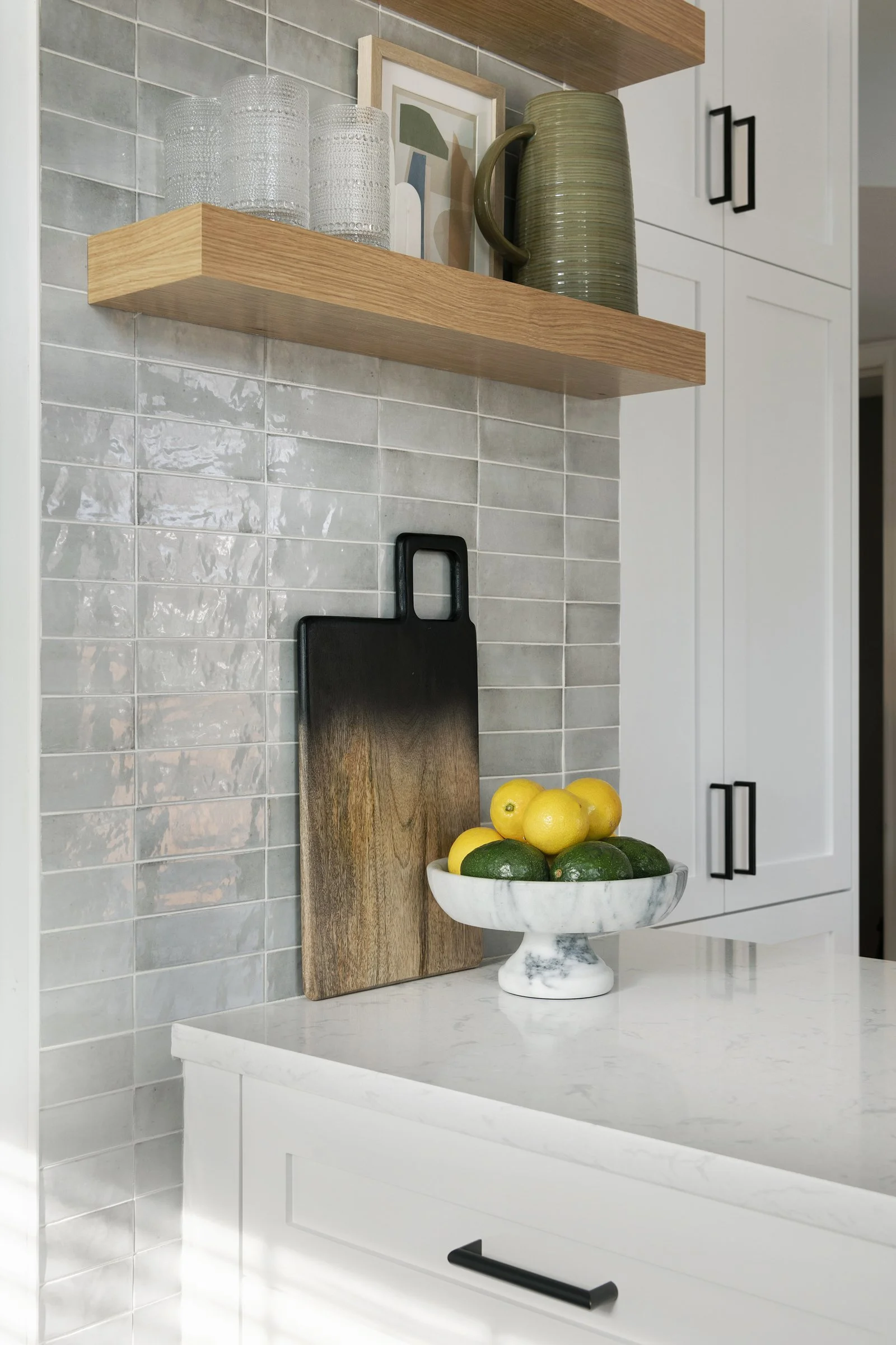 White kitchen cabinets with matte black hardware and a natural wood floating shelf positioned above a marble-tiled backsplash.