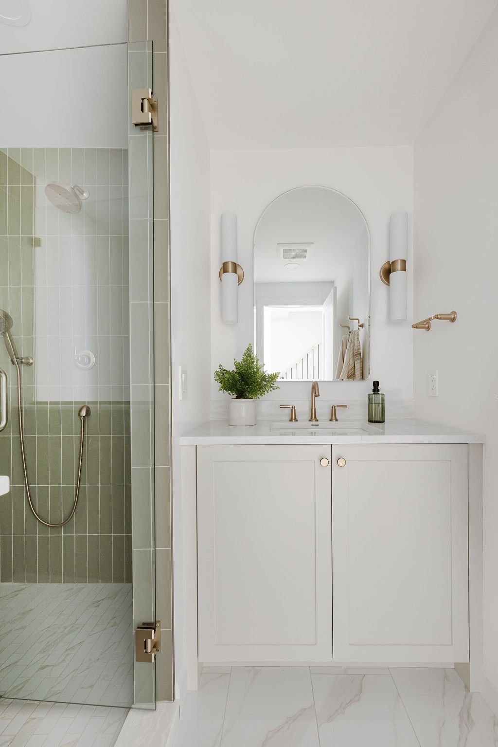 A full view of a modern bathroom featuring a white shaker-style vanity with gold knobs and a walk-in shower with sage green vertical tiles. The vanity is complemented by an arched mirror, gold fixtures, and symmetrical white sconces.