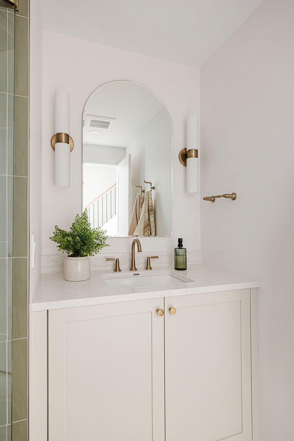 High-angle view of a custom white shaker vanity featuring gold hardware and a white quartz countertop. The setup includes an arched wall mirror framed by two modern brass-and-white cylinder sconces.