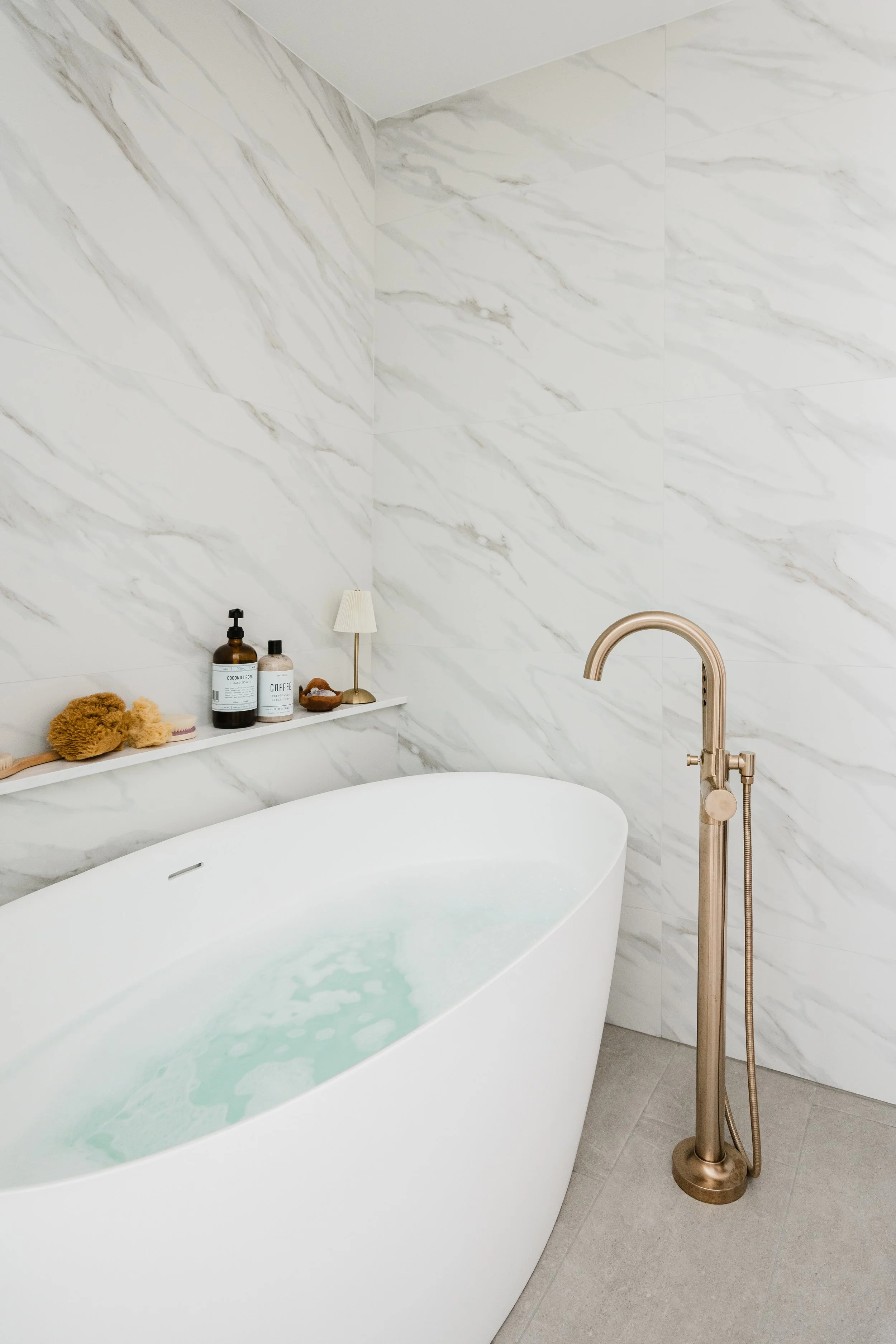 Freestanding tub with brass fixture and nearby light wood cabinets in a marble bathroom