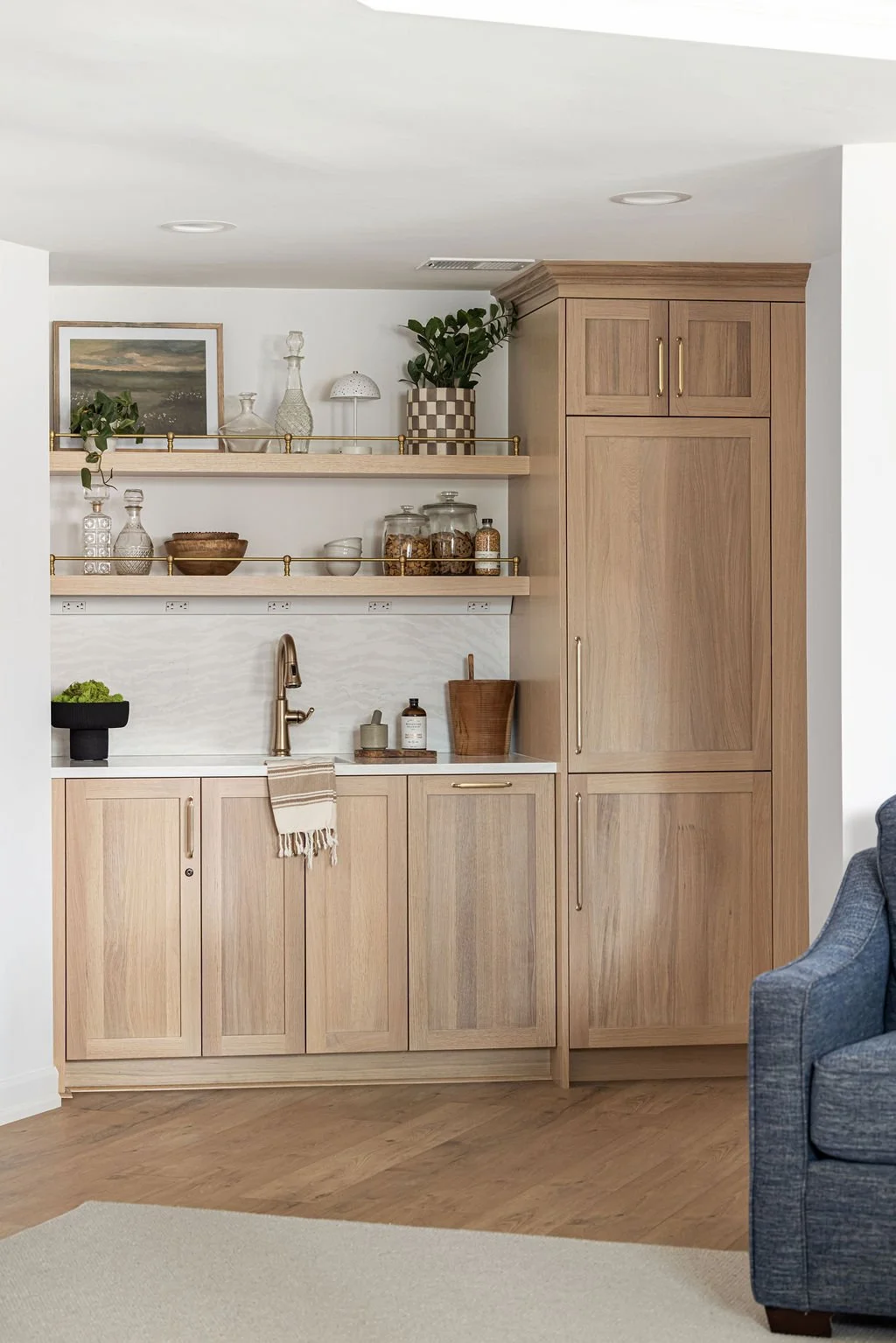 Custom light oak wet bar featuring flat-panel cabinetry and two floating shelves with brass gallery rails. A tall integrated cabinet unit stands to the right, complemented by a white marble backsplash and warm gold hardware.