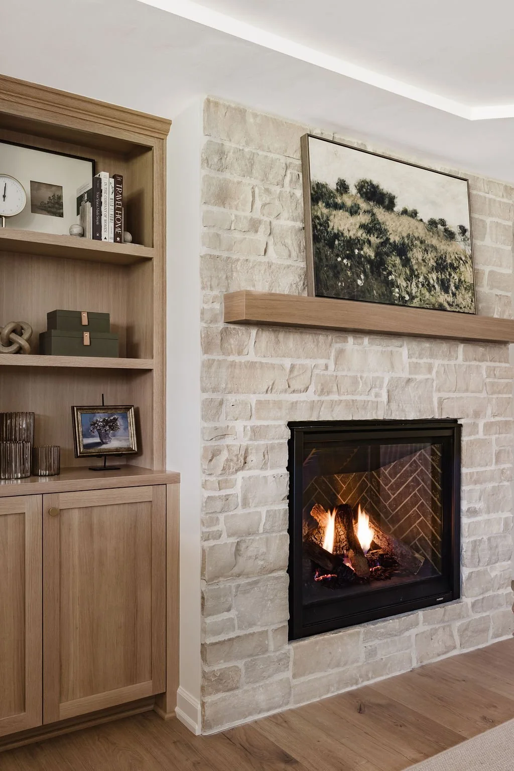 Modern living room featuring a floor-to-ceiling light stone fireplace with a natural wood mantel and a landscape painting above. To the left, a built-in light oak bookshelf with shaker-style lower cabinets displays curated decor, books, and framed ar