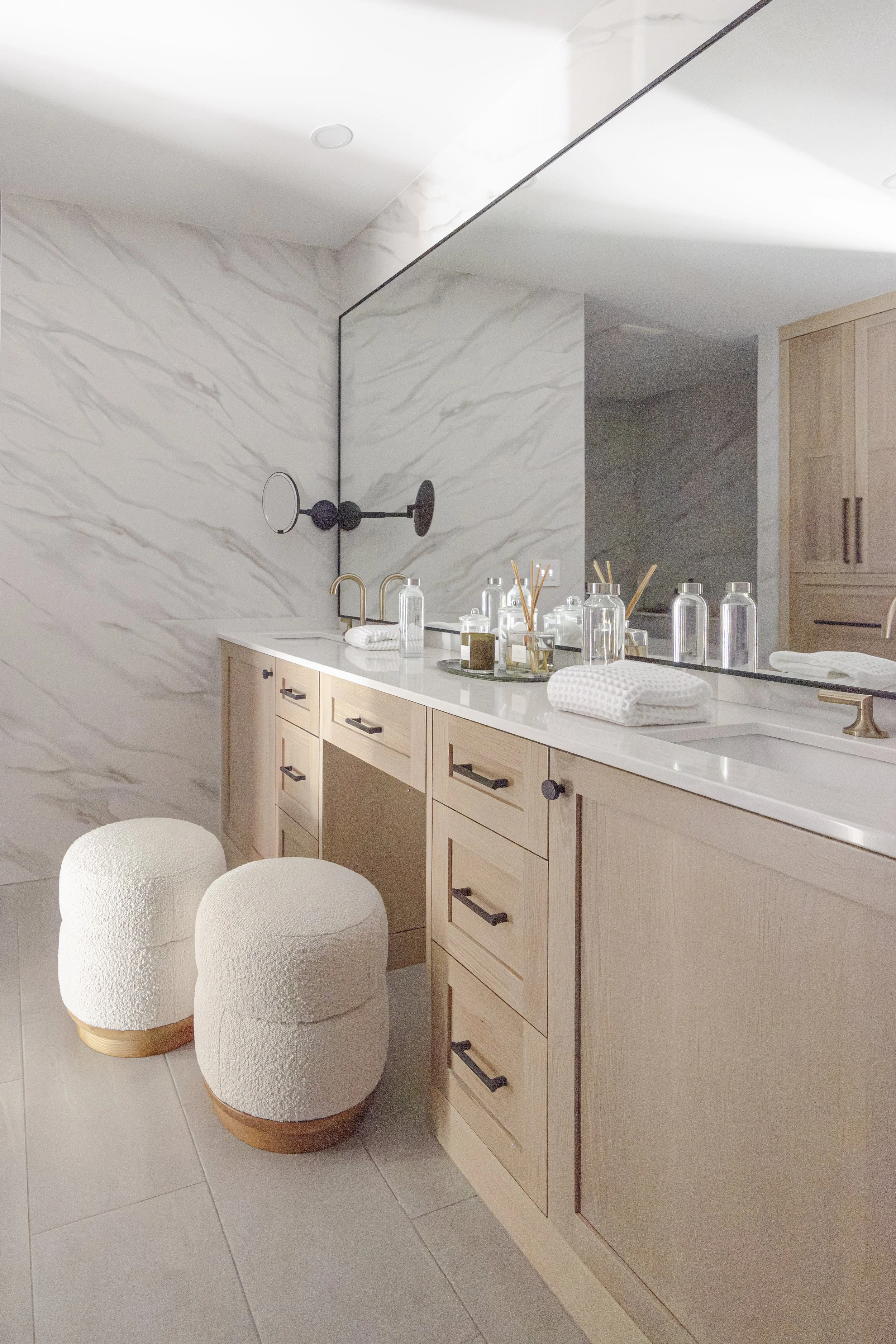 Light wood bathroom cabinets with marble backsplash, large mirror, and double vanity with upholstered stools.
