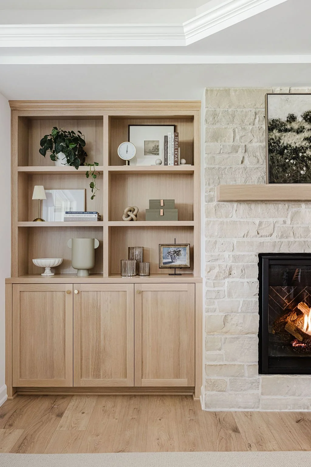 Custom light wood built-in cabinet with open shelving styled with decor, adjacent to a white stone fireplace in a modern living room.